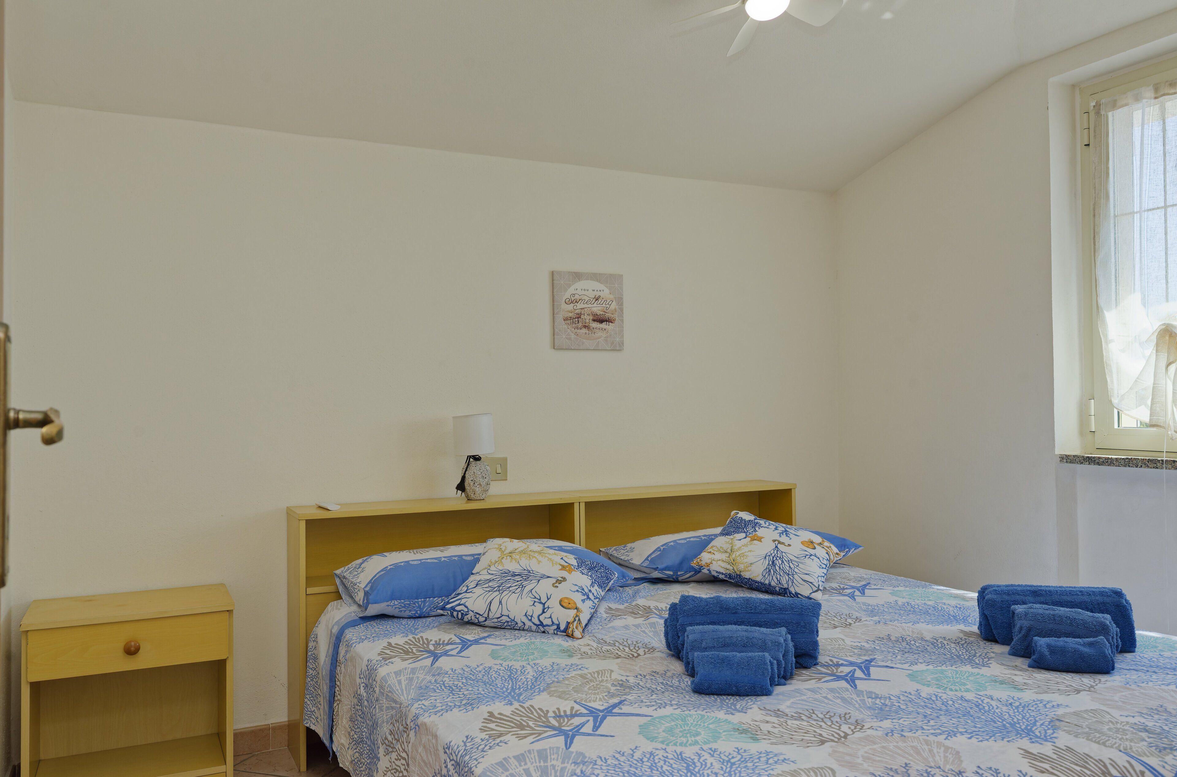 Un dormitorio con cama, mesita de noche, lámpara de pared y ventana con cortinas.