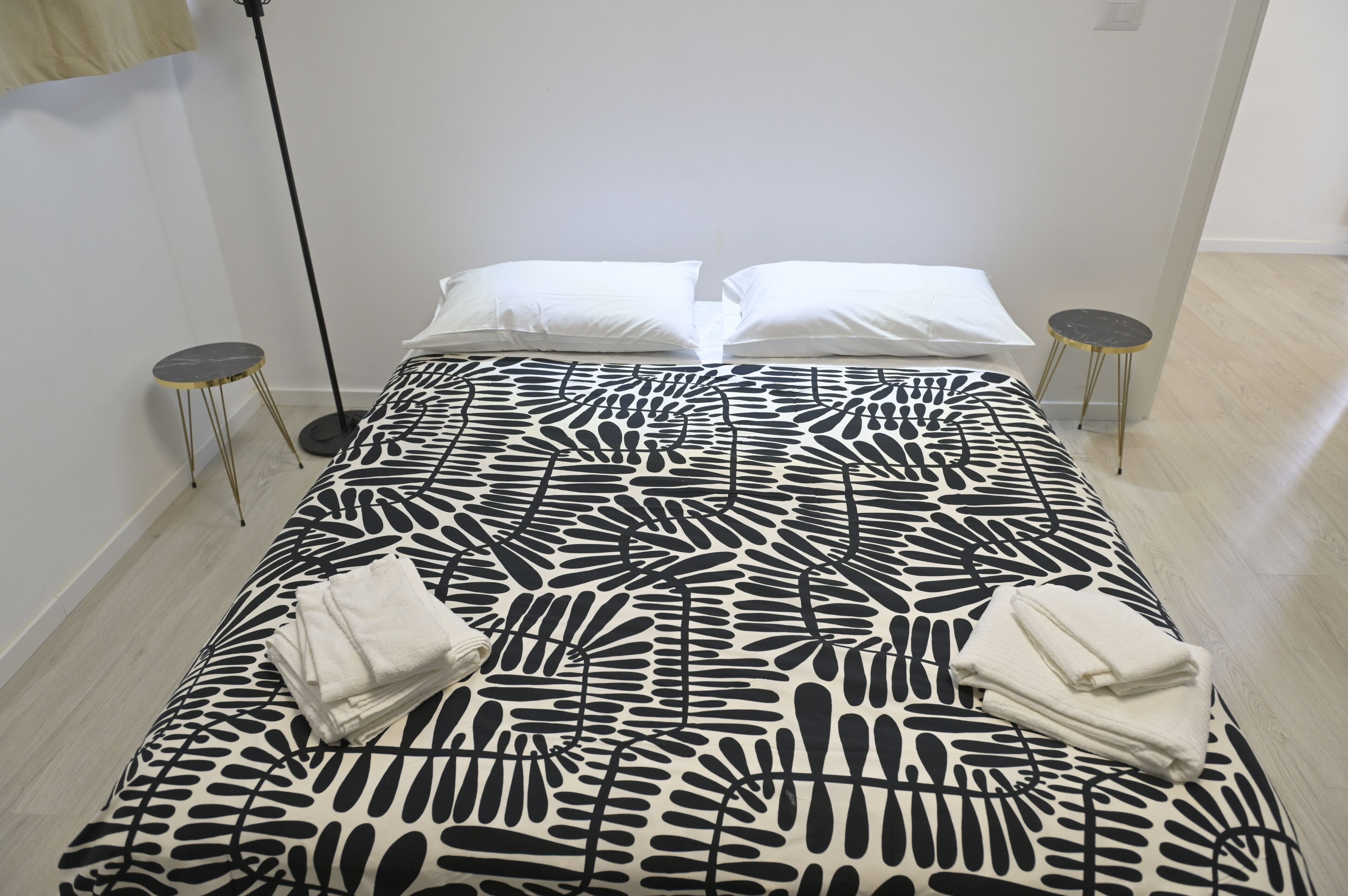 A neatly made bed with a black and white leaf-patterned bedspread, two white pillows, and two folded white towels. There are two small round tables with gold legs, one on each side of the bed.
