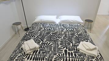 A neatly made bed with a black and white leaf-patterned bedspread, two white pillows, and two folded white towels. There are two small round tables with gold legs, one on each side of the bed.