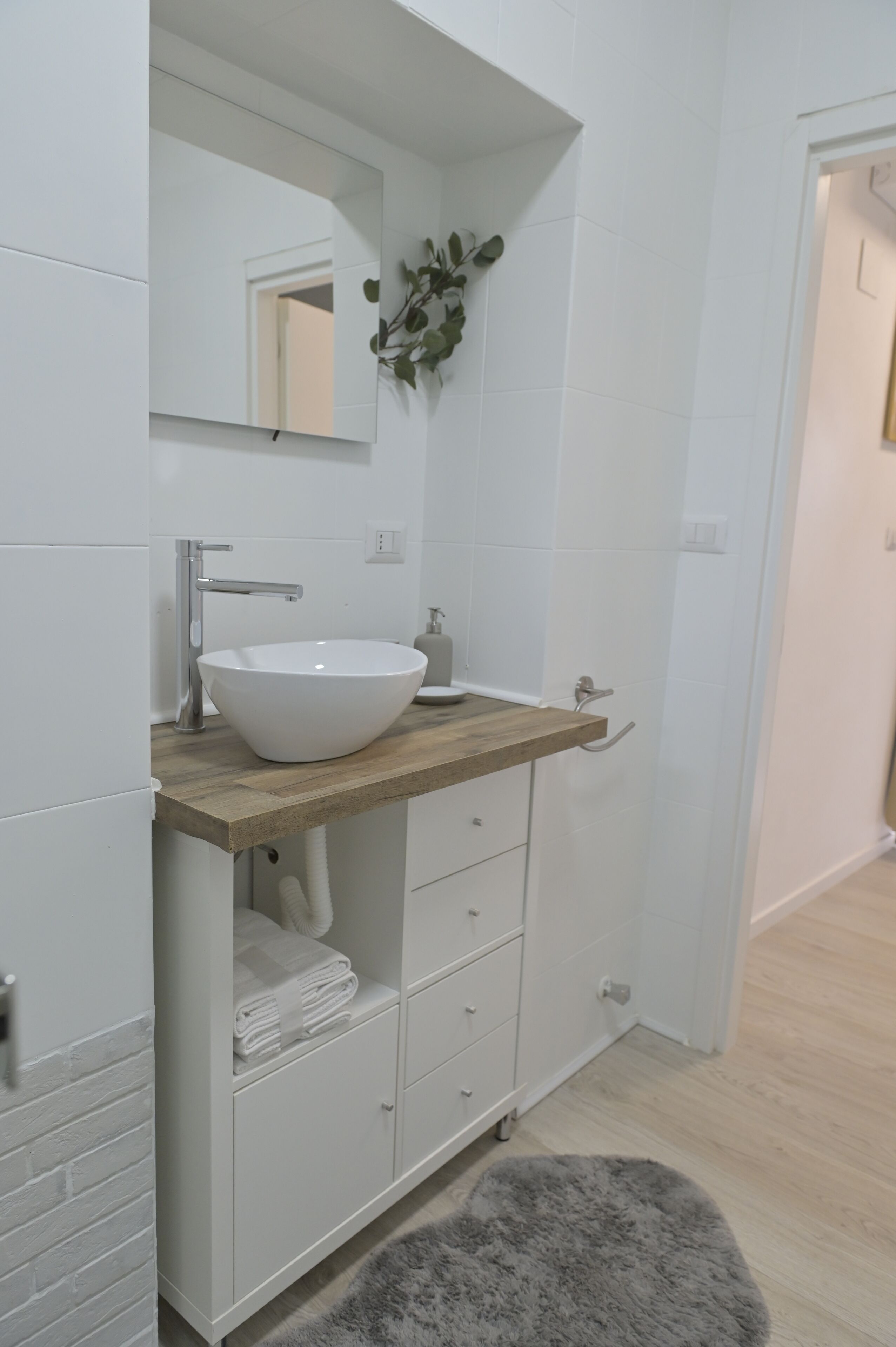 Een badkamer met een wit wastafel, een houten aanrechtblad en een spiegel. Er zijn witte kastjes onder het aanrechtblad en een grijs vloerkleed.