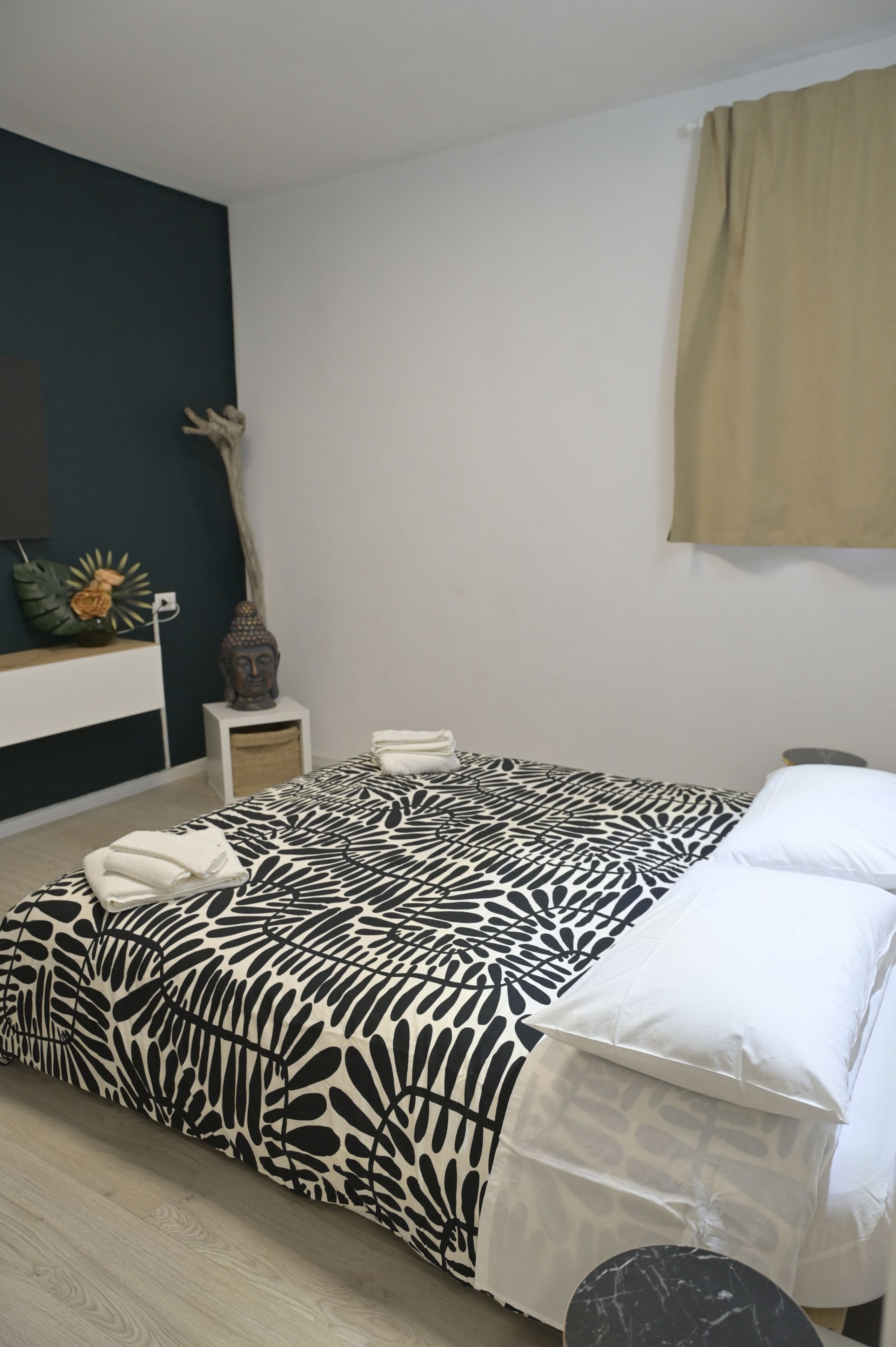 A bedroom with a patterned bedspread, a white bedside table with a vase, and a wall-mounted mirror.