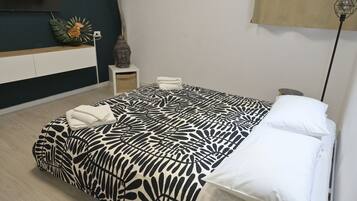 Una cama bien hecha con una colcha estampada, una mesita de noche con un jarrón y un televisor montado en la pared.