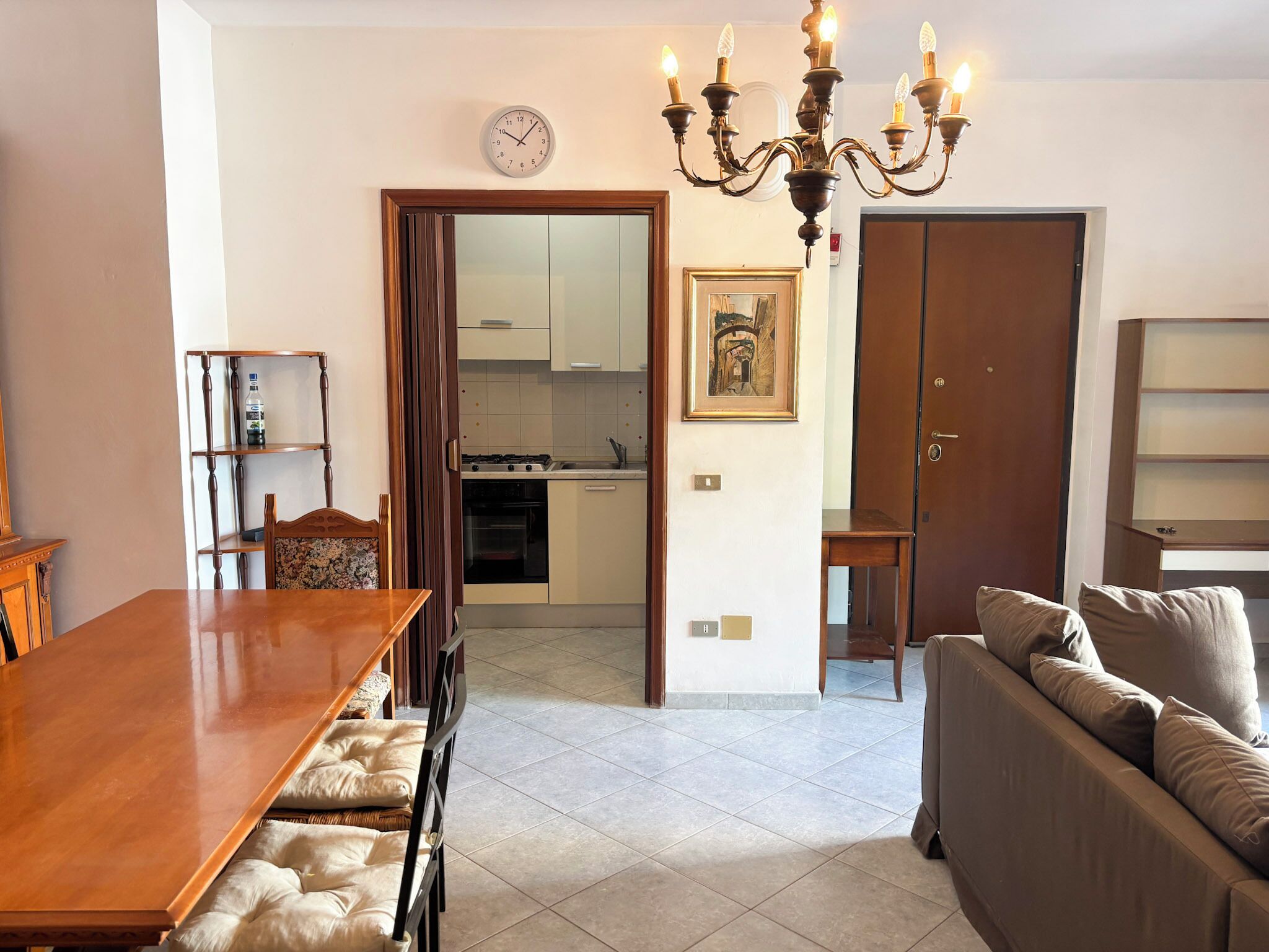 A dining area with a wooden table and chairs, a sofa, a chandelier, a kitchen in the background, and a door leading to another room.