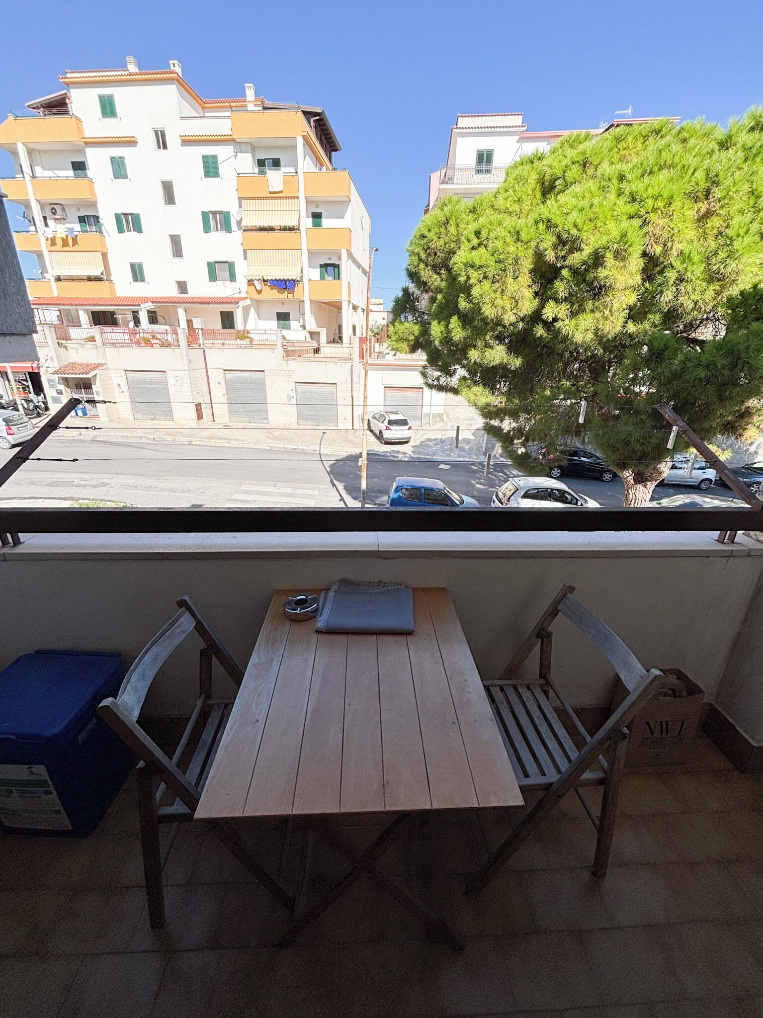 Uma varanda com mesa de madeira e duas cadeiras, com vista para uma rua com carros estacionados e edifícios.