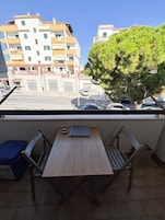 Un balcone con un tavolo di legno e due sedie, affacciato su una strada con auto parcheggiate e palazzi.