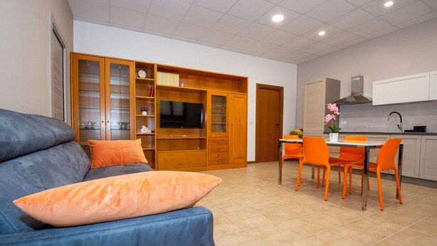 Ein modernes Wohnzimmer mit einem blauen Sofa, orangefarbenen Kissen und einem Fernseher. Es gibt eine Essecke mit orangefarbenen Stühlen und eine Küche im Hintergrund.