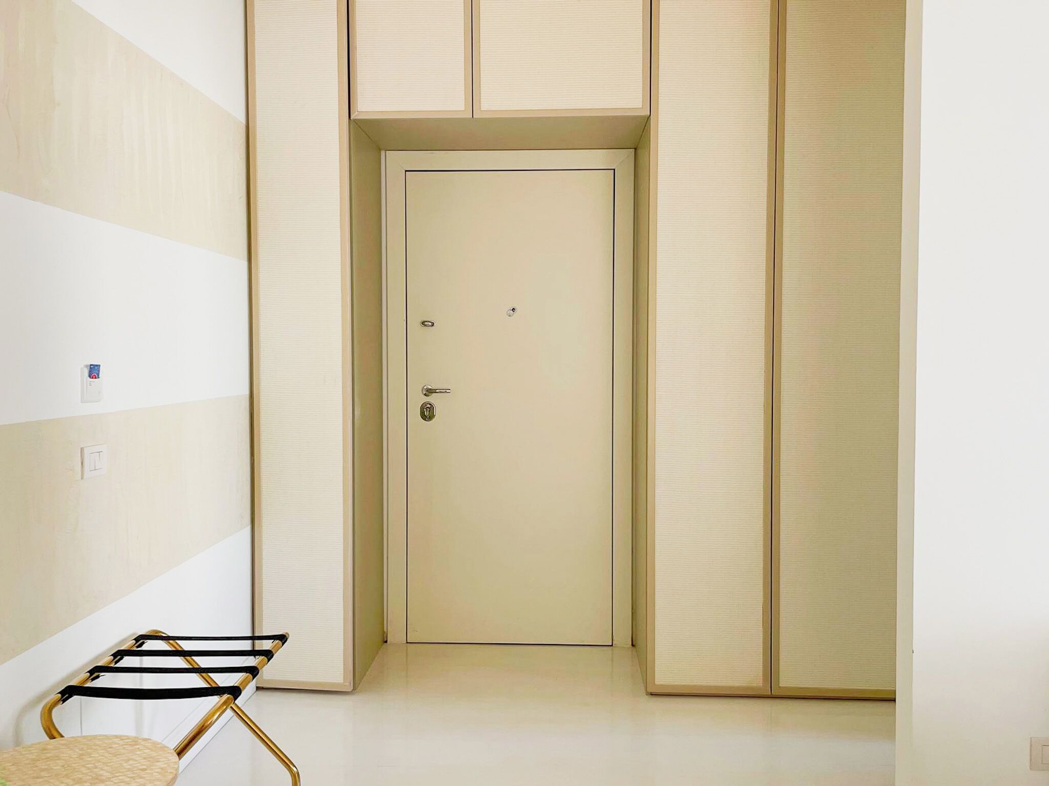 Un pasillo estrecho con una puerta color beige, una silla con marco dorado y un interruptor de luz en la pared.