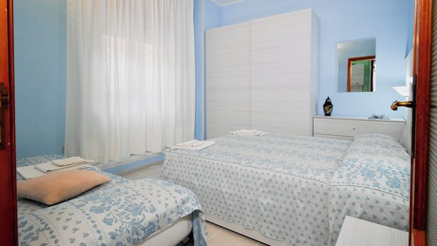 A bedroom with two beds, a white wardrobe, a bedside table with a lamp, and a window with curtains. - Casa Ida (San Giovanni Rotondo)