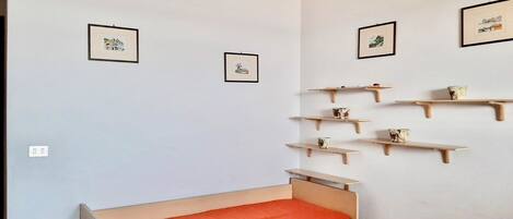 A small, single-bed room with a wooden bed and a red mattress. There are two framed pictures on the wall, and a series of wooden shelves with decorative items.