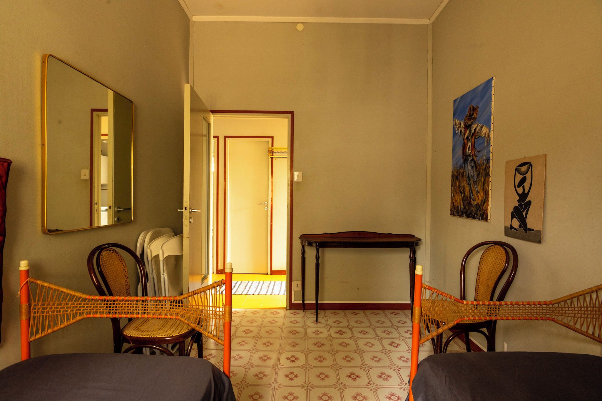 Um quarto com duas cadeiras de vime, uma mesinha, um espelho e obras de arte nas paredes.