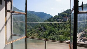Blick durch ein Fenster mit Metallrahmen auf eine gebirgige Landschaft mit grünen Flächen und einigen Gebäuden.