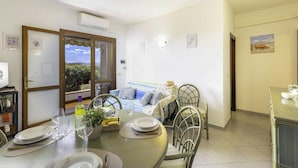 Zona pranzo con tavolo apparecchiato per due, divano con cuscini e vista attraverso una finestra.