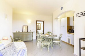A dining area with a round table and chairs, a kitchen area with cabinets and appliances, and a bed with a hat and blanket. - Venere (Agrustos)