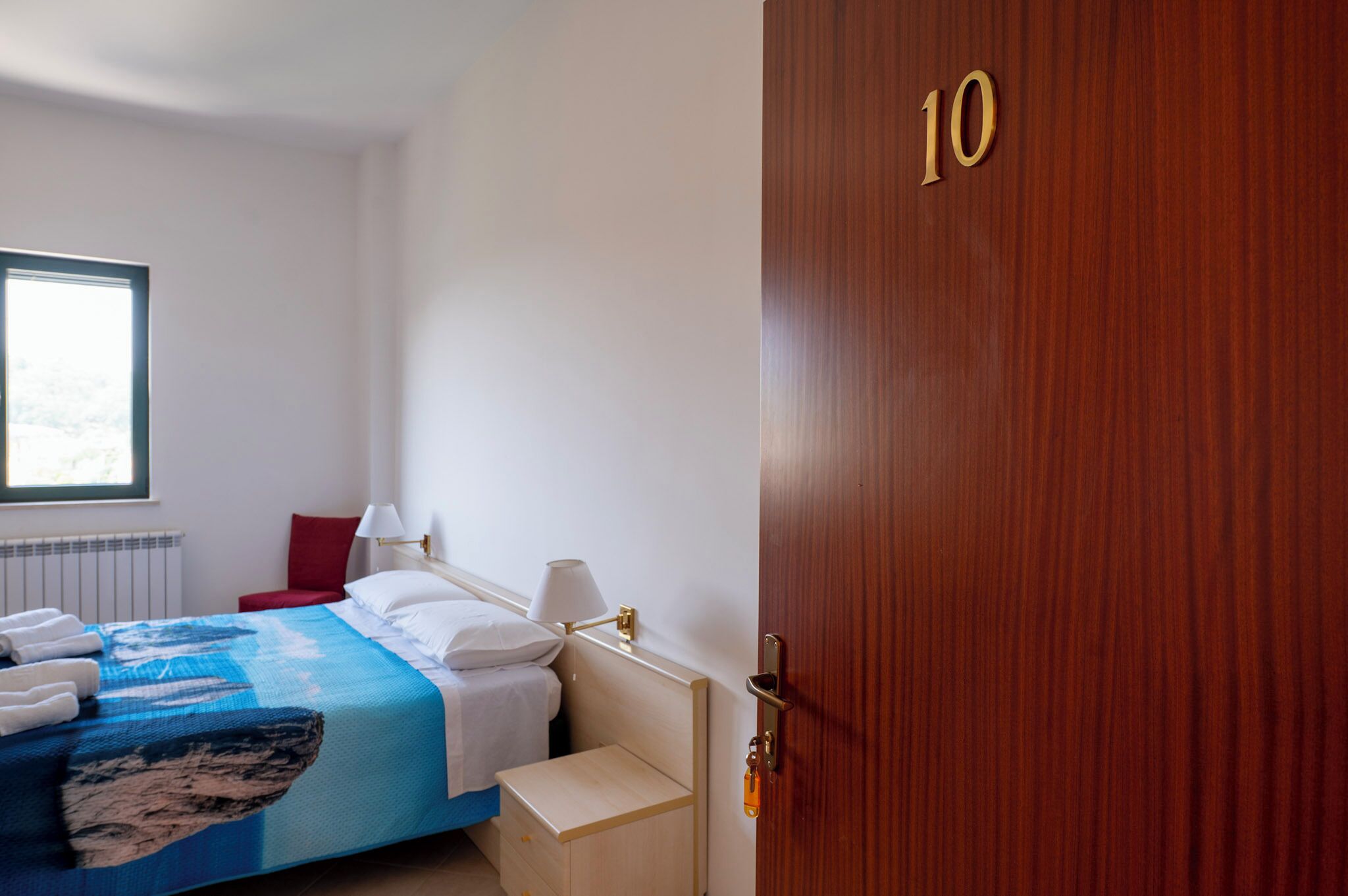 Une chambre d’hôtel avec un lit, une fenêtre, une porte portant le numéro 10 et une table de chevet.