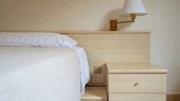 Un letto rifatto con un copriletto bianco e cuscini, una lampada da comodino e un comodino in legno con cassetti.