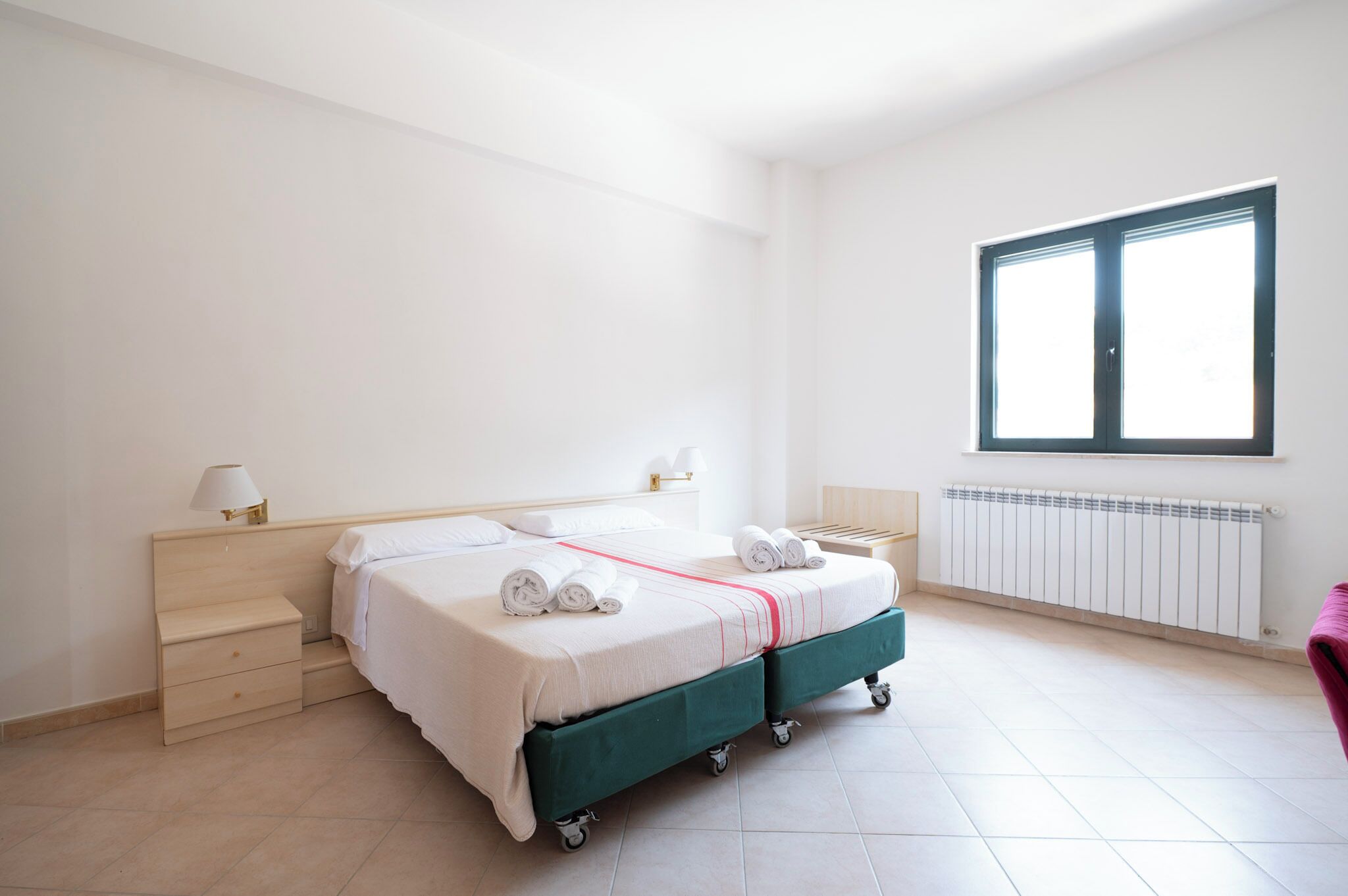 A hotel room with a double bed, a nightstand, a lamp, a window, and a radiator.