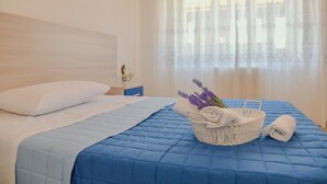 A neatly made bed with a blue quilt, a white pillow, a white basket with towels and lavender, and a nightstand with a lamp. - Le Sciosce (San Giorgio Lucano)