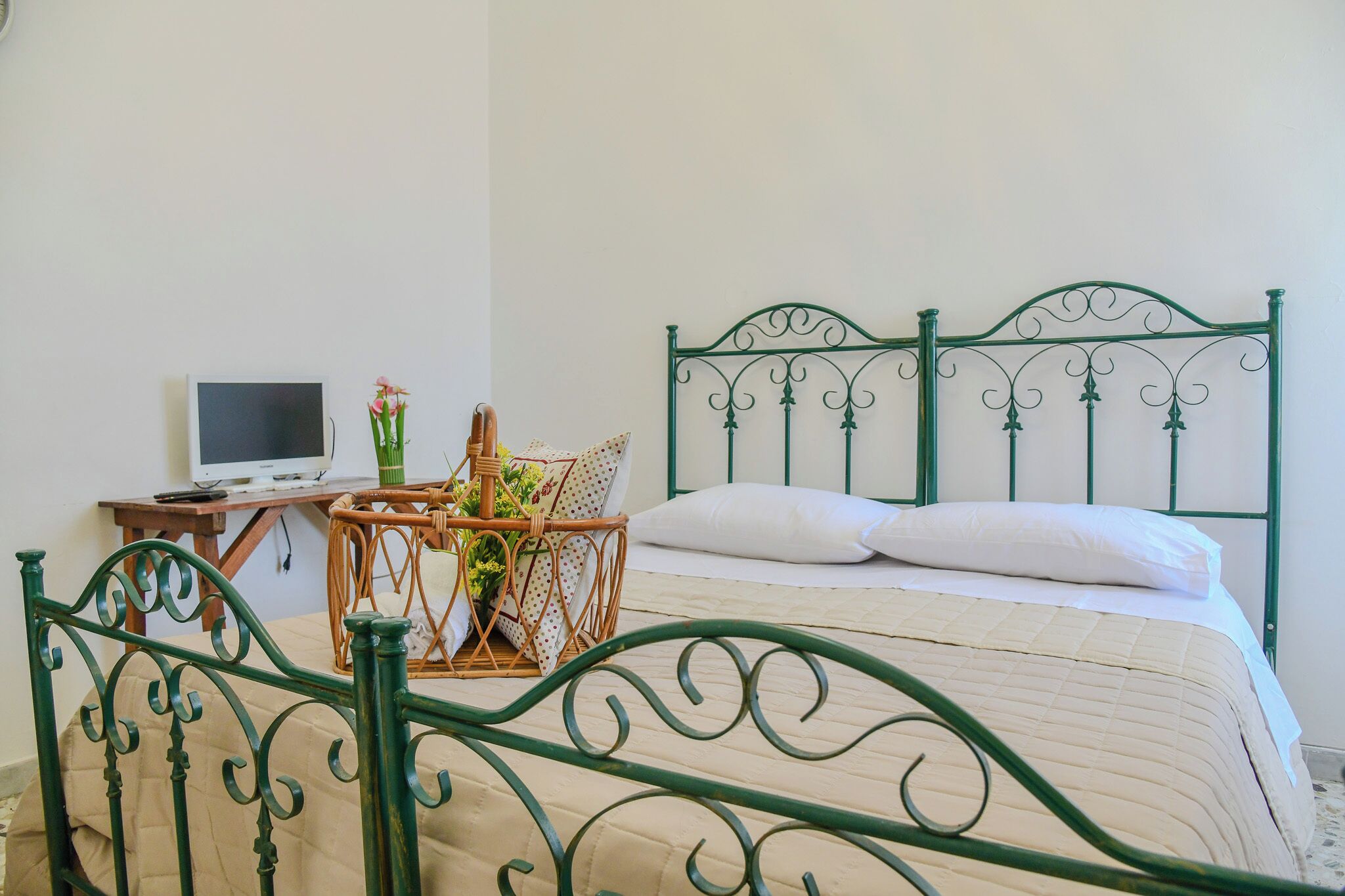 Una cama con cabecera de metal verde, una mesita de noche con una computadora portátil y una silla de mimbre con un jarrón de flores.