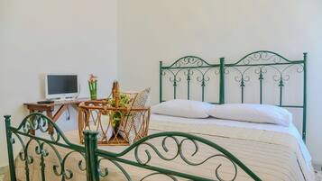 Una cama con cabecera de metal verde, una mesita de noche con una computadora portátil y una silla de mimbre con un jarrón de flores.