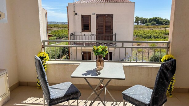 A balcony with two chairs and a table, a vase of flowers, and a view of a building with a tiled roof. - Sofia Kite Apartment (Marsala)