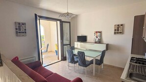 A compact living space with a red sofa, a glass dining table with chairs, a kitchenette, and a sliding glass door leading to an outdoor area. - Sofia Kite Apartment (Marsala)