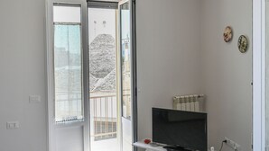 A modern living room with a flat-screen TV on a wooden stand, a glass table with decorative items, and a balcony with a view. - Il Rifugio Di Eva (Alberobello)