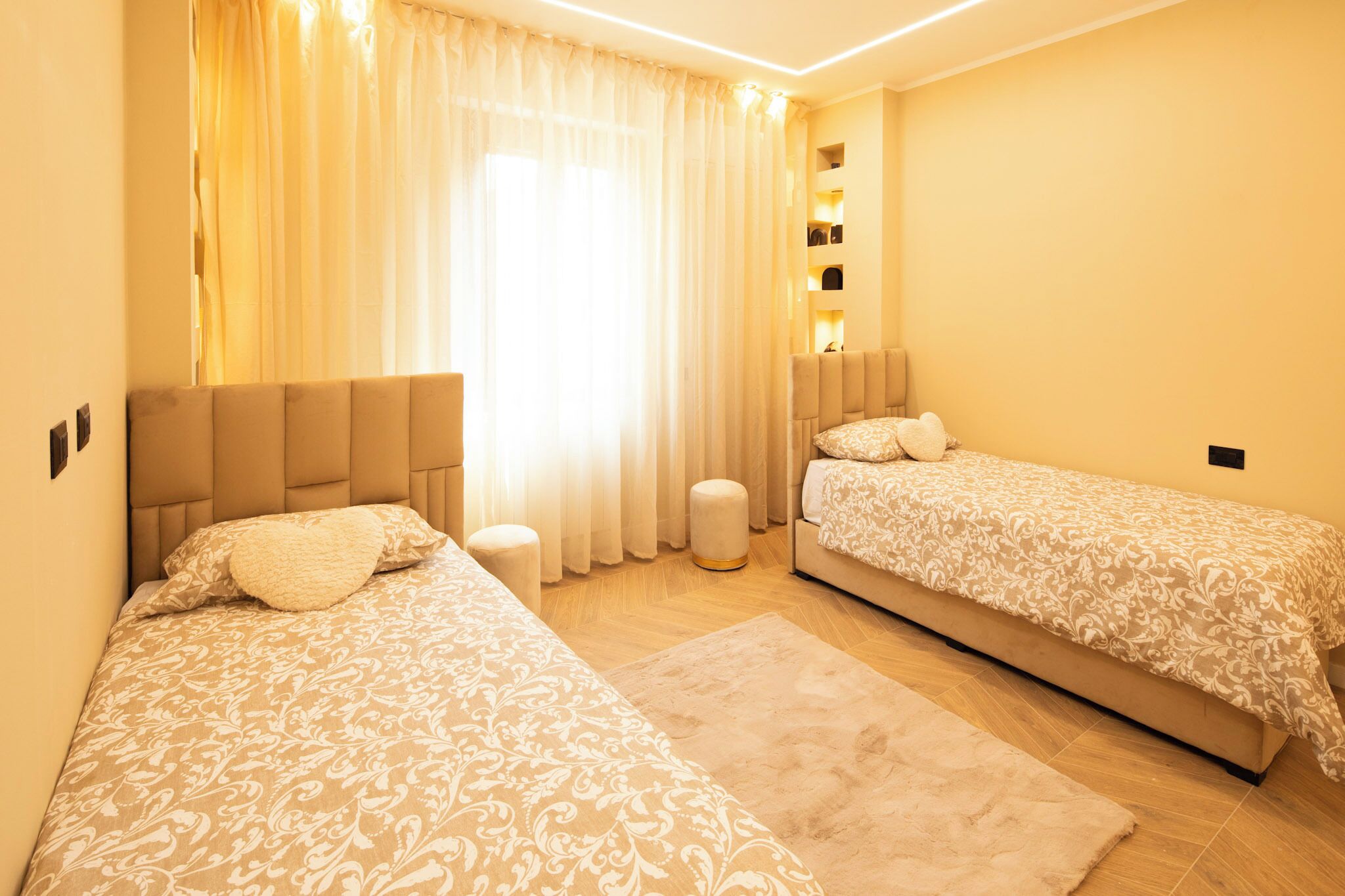 A bedroom with two beds, a large window with sheer curtains, a built-in shelf with decorative items, and a small ottoman.