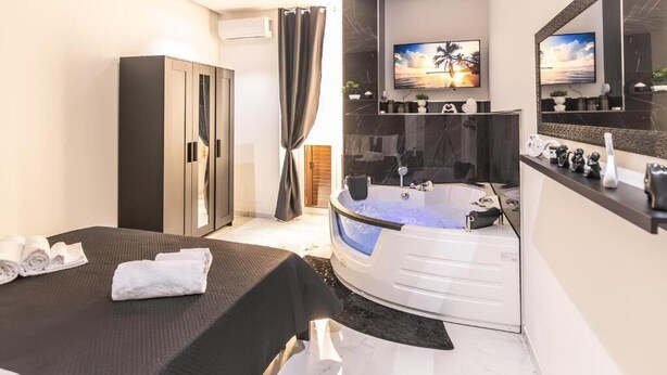 A modern bathroom with a large bathtub, a vanity with a mirror, and a television mounted above the sink. - Amour (Rosolini)