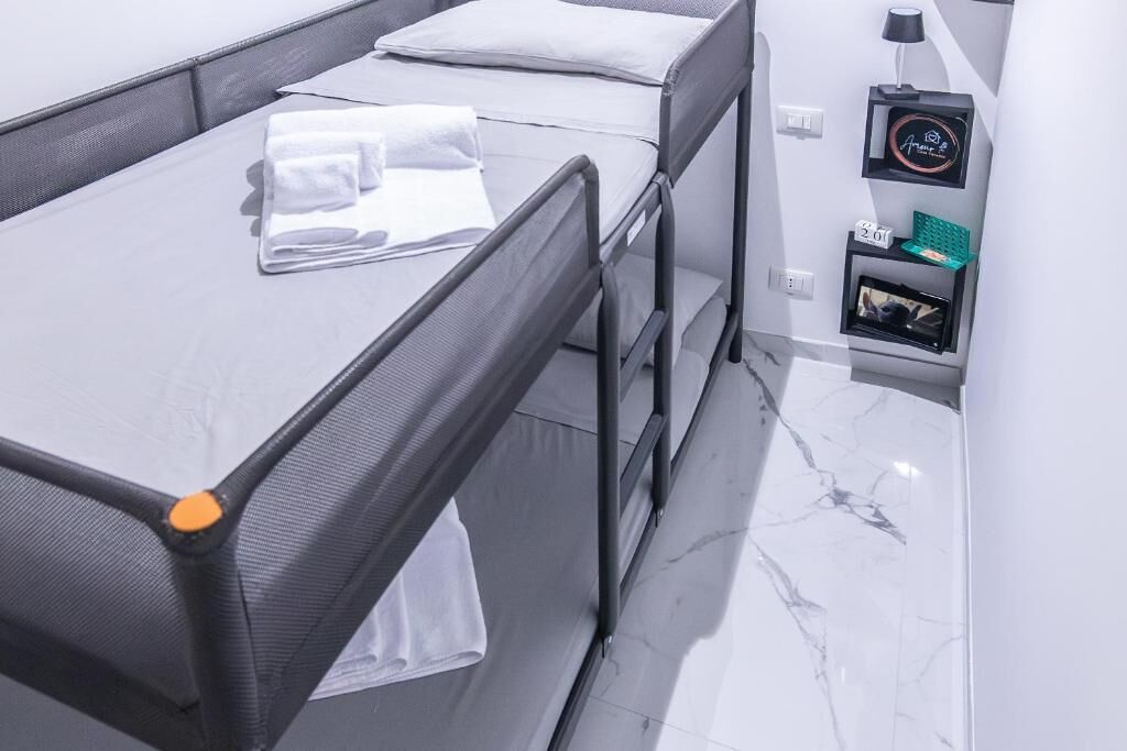 Habitación de hotel con una cama, una mesita de noche con lámpara y una estantería en la pared con un reloj y un marco de fotos.