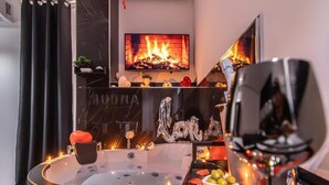 A modern bathroom with a freestanding bathtub, a mirror, and a wall-mounted TV displaying a fireplace. - Amour (Rosolini)