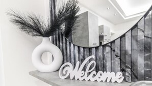 A modern interior with a 'Welcome' sign, a white vase with decorative branches, and a curved mirror reflecting the room. - Amour (Rosolini)