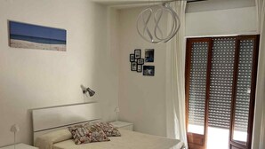 A bedroom with a bed, a nightstand, a wall-mounted lamp, a ceiling light fixture, a window with blinds, and a patterned floor. - Perla Sul Mare (Vibo Marina)