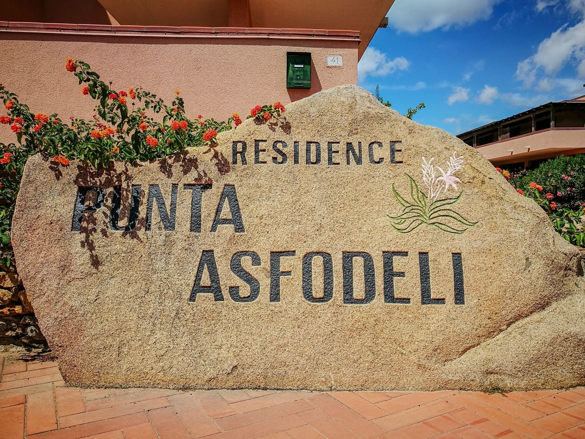 Une plaque en pierre portant l’inscription « RÉSIDENCE PUNTA ASFODELI », entourée de plantes et de fleurs.