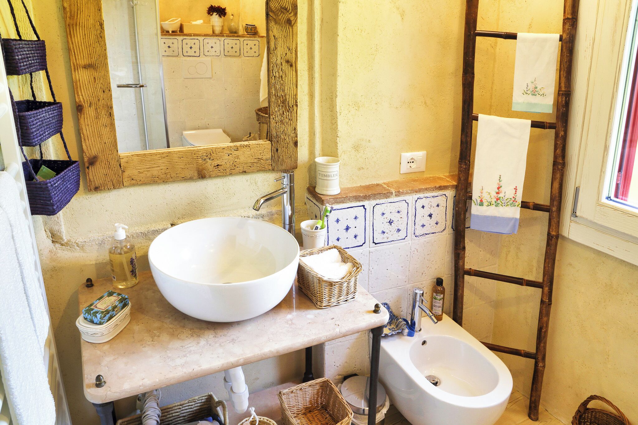 A bathroom with a large white basin, a wooden shelf with baskets, a wooden ladder, and a window with a towel hanging.