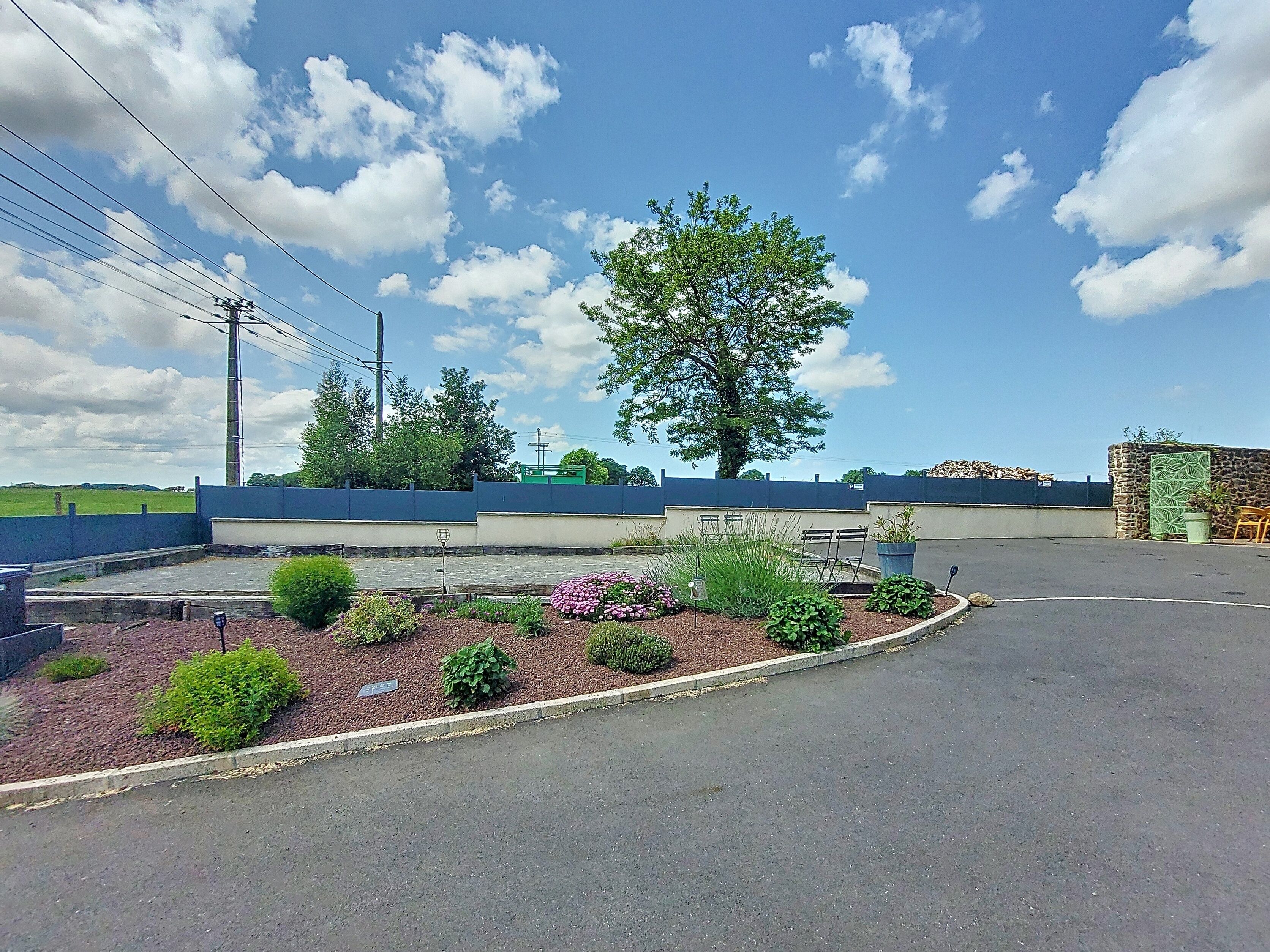 A parking lot with a landscaped garden area, a large tree, and a building with a blue roof in the background.