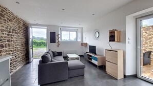 A modern living room with a grey sofa, a TV, and a wooden cabinet. There is a stone wall on one side and a large window providing natural light.