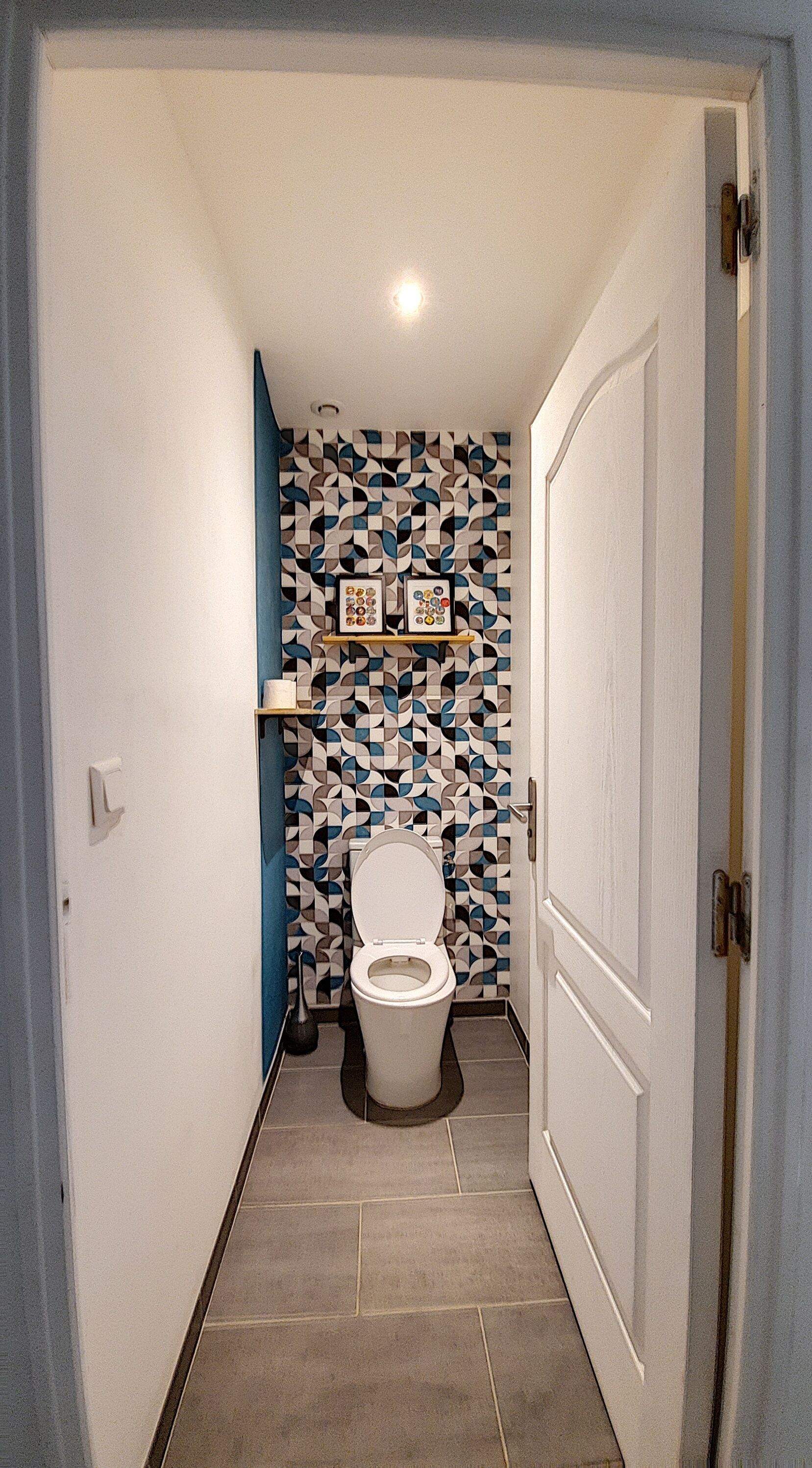 Ein schmaler Flur führt zu einem Badezimmer mit Toilette, einem gemusterten Wandbelag und einer Tür auf der rechten Seite.