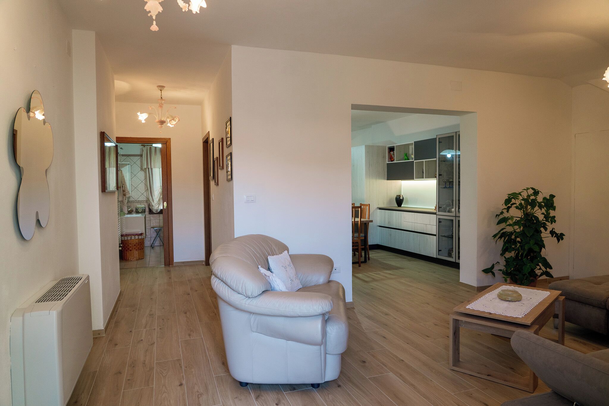 Una sala moderna con un sofá de cuero color beige, una mesita de centro de madera y una planta en maceta. Se observa una cocina al fondo con armarios blancos y una zona de comedor.