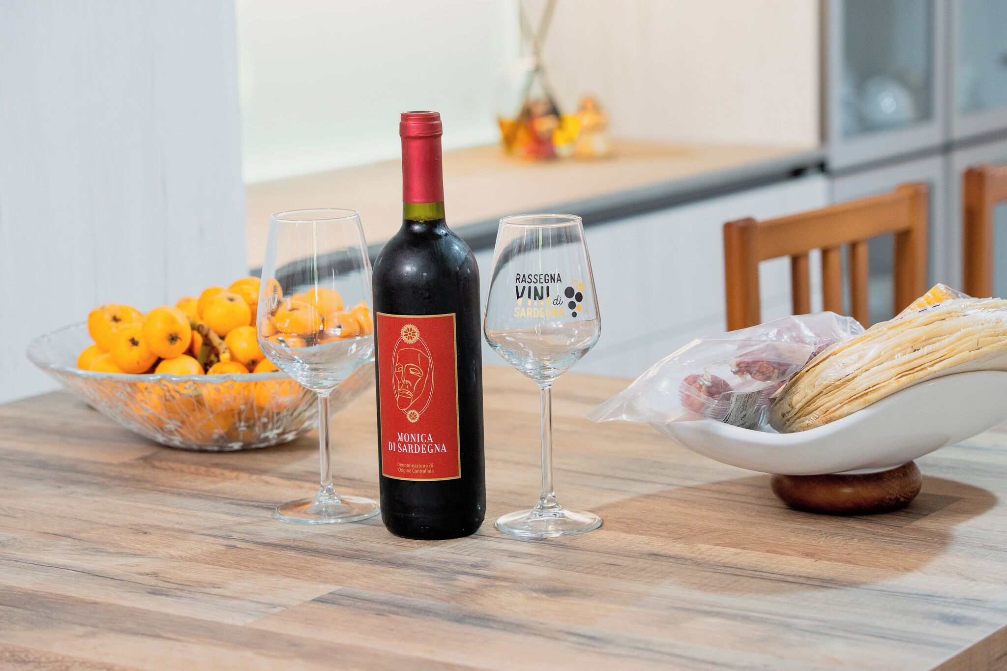 A dining table with a bottle of wine, two glasses, a bowl of oranges, and a bowl of bread.