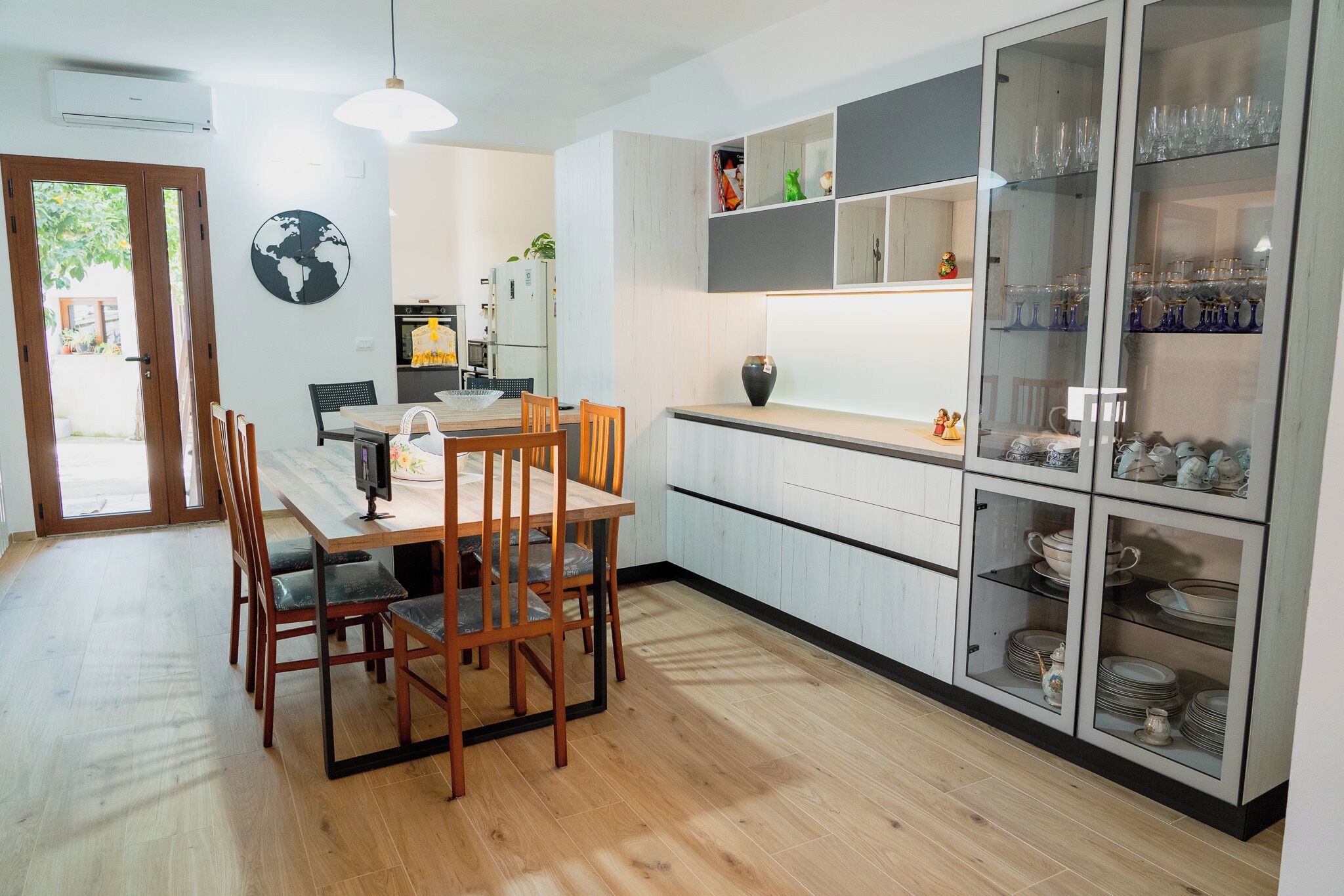 Uma cozinha moderna com soalho de madeira, uma mesa de jantar com cadeiras e armários embutidos com portas de vidro que exibem louças e utensílios de cozinha.