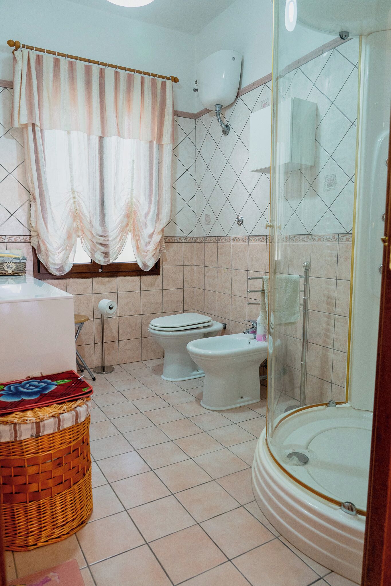 A bathroom with a bathtub, two toilets, a shower, a wicker basket, and a window with curtains.