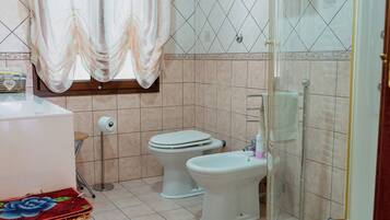 A bathroom with a bathtub, two toilets, a shower, a wicker basket, and a window with curtains.