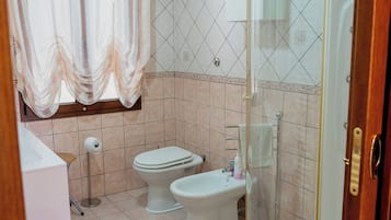 A bathroom with two toilets, a shower, a toilet paper holder, and a basket.