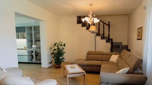 A living room with a sofa, armchair, and a coffee table. A staircase with a wooden railing is visible in the background.