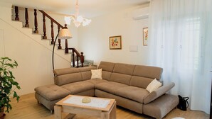 A living room with a brown sectional sofa, a wooden coffee table, a staircase with wooden railings, and a chandelier.
