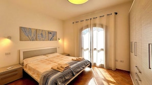 A bedroom with a bed, a nightstand, a wall-mounted light, a window with curtains, and a door. - Casa A.Danieli (Olbia)