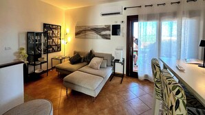 A living room with a sofa, a coffee table, a side table, a bookshelf, and a large window with curtains. - Casa A.Danieli (Olbia)