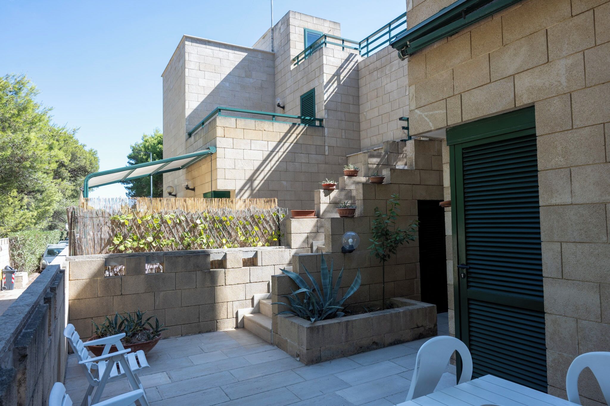 Un patio exterior moderno con muros de piedra, plantas en macetas y mobiliario blanco.
