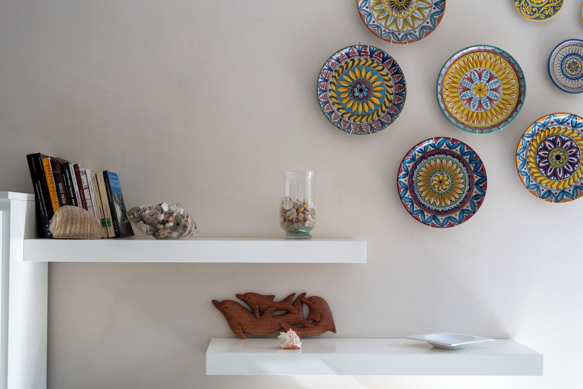 A room with a white wall, a shelf with books, decorative plates, and a wooden dolphin sculpture.