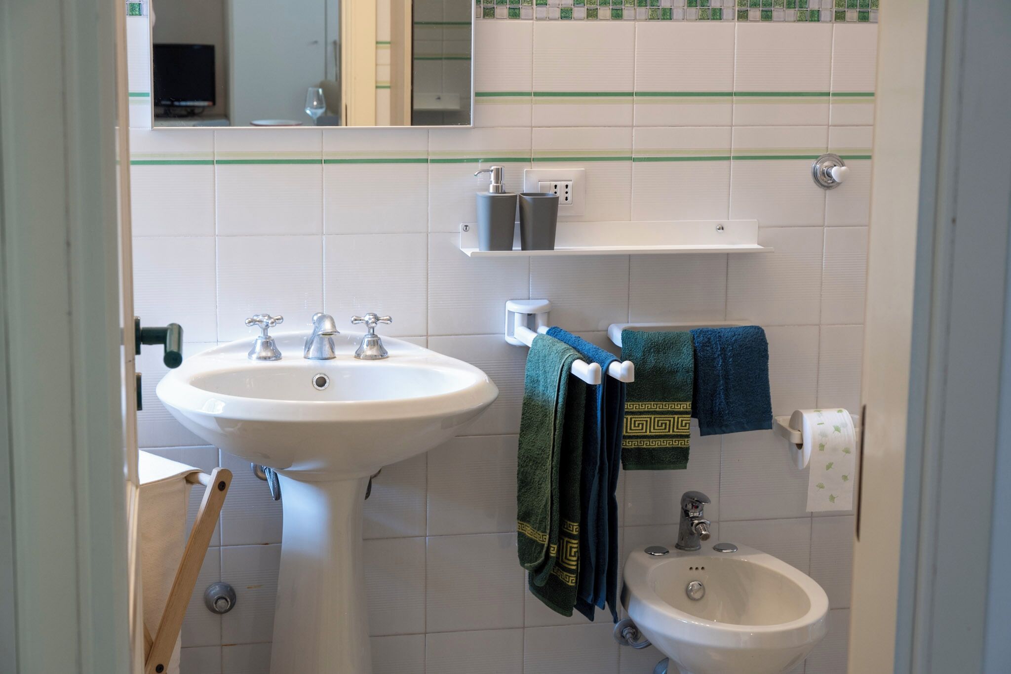 A bathroom with a pedestal sink, a smaller sink, and a bidet. There are towels hanging on a rack and a shelf with toiletries.